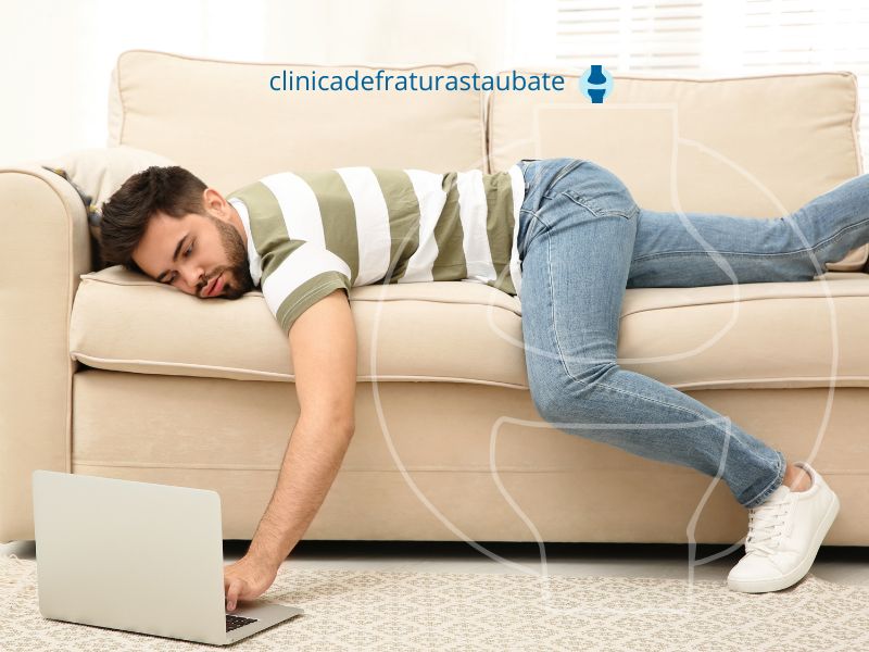 Homem sedentário deitado no sofá e mexendo no laptop.